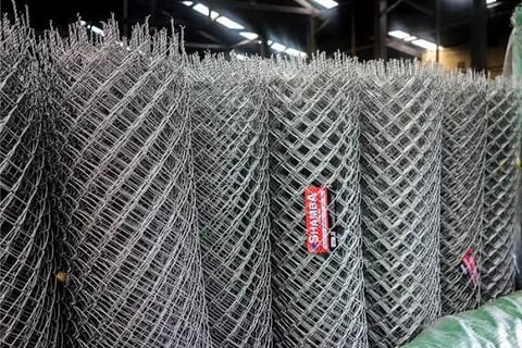 Rolls of metal chain-link fencing stacked in an industrial warehouse with fluorescent lighting